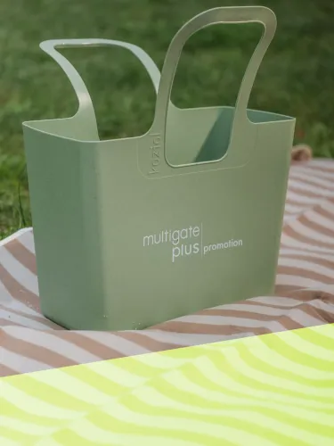 Grüner Kunststoffbeutel mit Logo und Text "multigate plus promotion" auf einer Picknickdecke.